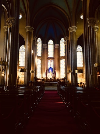 A grand and ornate church interior featuring tall, majestic columns and a series of tall arched stained-glass windows that allow light to create a warm glow inside. The altar is illuminated with a blue light centerpiece, and the aisle is covered with a dark red carpet. The atmosphere is serene and reverent.