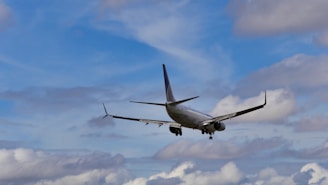 A commercial airplane is in flight against a backdrop of a partly cloudy sky. The aircraft is viewed from the rear, revealing its wings, tail fin, and landing gear. The sky features a mix of blue and white clouds, with varying shades of both colors.