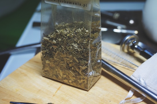 A clear plastic bag filled with dried green leaves sits on a wooden cutting board. Surrounding the bag are kitchen utensils and tools in the background, including a metal straw, a white fabric pouch, and metallic items creating a kitchen-like setting.