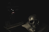 A dimly lit scene featuring a metallic skull resting on an open book. The setting is dark, with a single candle providing a faint glow, casting shadows over the objects nearby. The atmosphere is eerie and mysterious.