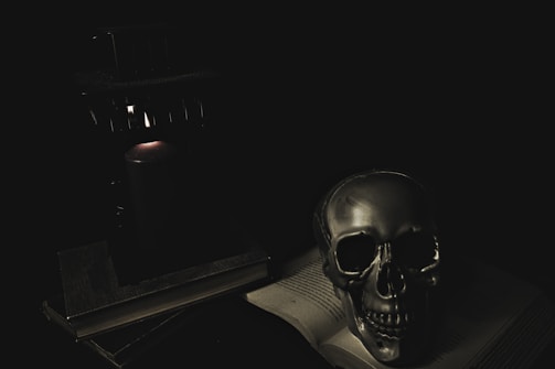 A dimly lit scene featuring a metallic skull resting on an open book. The setting is dark, with a single candle providing a faint glow, casting shadows over the objects nearby. The atmosphere is eerie and mysterious.
