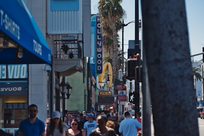 A busy urban street scene with a crowd of people walking near iconic Hollywood landmarks. Visible signage includes a vertical 'HOLLYWOOD' sign and a large yellow arch, indicating a recognizable fast-food restaurant. Palm trees line the street, and various shops are seen on either side of the crowded sidewalk.