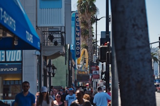 A busy urban street scene with a crowd of people walking near iconic Hollywood landmarks. Visible signage includes a vertical 'HOLLYWOOD' sign and a large yellow arch, indicating a recognizable fast-food restaurant. Palm trees line the street, and various shops are seen on either side of the crowded sidewalk.