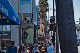 A busy urban street scene with a crowd of people walking near iconic Hollywood landmarks. Visible signage includes a vertical 'HOLLYWOOD' sign and a large yellow arch, indicating a recognizable fast-food restaurant. Palm trees line the street, and various shops are seen on either side of the crowded sidewalk.