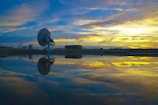 A large satellite dish is situated near a body of water, reflecting its image. The sky is filled with dramatic clouds, and the colors of sunrise or sunset are reflected in the water. There is a building and some utility poles in the background.