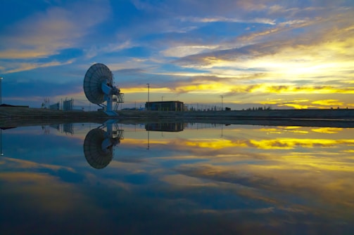 A large satellite dish is situated near a body of water, reflecting its image. The sky is filled with dramatic clouds, and the colors of sunrise or sunset are reflected in the water. There is a building and some utility poles in the background.