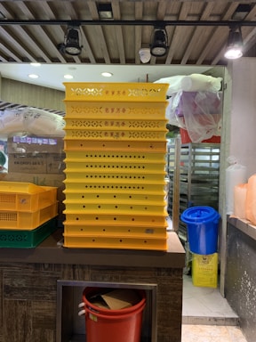 A stack of yellow perforated trays with red Chinese characters is positioned on a counter. Next to it, a green and yellow bin is visible with a blue and a red bin nearby. The setting appears to be a kitchen with hanging overhead lights and various supplies in the background.