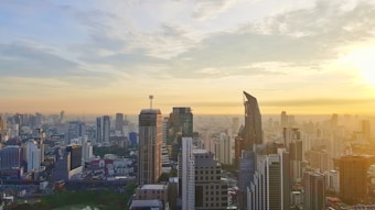A panoramic cityscape depicting a bustling urban environment with towering skyscrapers under a partially cloudy sky. The sun is setting or rising on the horizon, casting a warm glow over the buildings.
