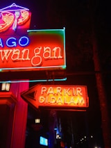 Bright neon signs glow vibrantly against a dark background, featuring red, blue, and green text. A prominent arrow points to the right with the words 'PARKIR DI DALAM', indicating parking inside. Various elements of the signs, including symbols like a fork and spoon above, suggest a restaurant or dining establishment.