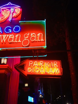 Bright neon signs glow vibrantly against a dark background, featuring red, blue, and green text. A prominent arrow points to the right with the words 'PARKIR DI DALAM', indicating parking inside. Various elements of the signs, including symbols like a fork and spoon above, suggest a restaurant or dining establishment.