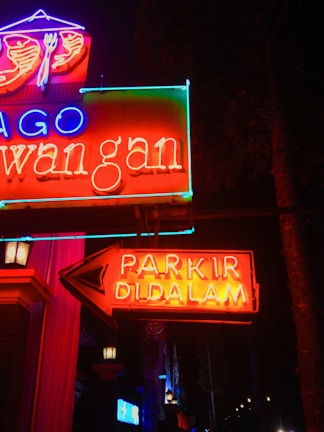 Bright neon signs glow vibrantly against a dark background, featuring red, blue, and green text. A prominent arrow points to the right with the words 'PARKIR DI DALAM', indicating parking inside. Various elements of the signs, including symbols like a fork and spoon above, suggest a restaurant or dining establishment.