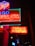Bright neon signs glow vibrantly against a dark background, featuring red, blue, and green text. A prominent arrow points to the right with the words 'PARKIR DI DALAM', indicating parking inside. Various elements of the signs, including symbols like a fork and spoon above, suggest a restaurant or dining establishment.