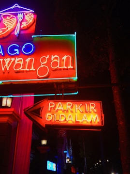 Bright neon signs glow vibrantly against a dark background, featuring red, blue, and green text. A prominent arrow points to the right with the words 'PARKIR DI DALAM', indicating parking inside. Various elements of the signs, including symbols like a fork and spoon above, suggest a restaurant or dining establishment.