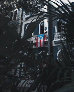 A building with dark blue and white walls, featuring ornate white balconies. A Puerto Rican flag hangs prominently from the balcony. Lush green foliage frames the image, adding a sense of seclusion and tranquility.