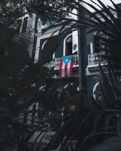 A building with dark blue and white walls, featuring ornate white balconies. A Puerto Rican flag hangs prominently from the balcony. Lush green foliage frames the image, adding a sense of seclusion and tranquility.