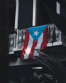 A Puerto Rican flag hangs from a wrought iron balcony, with intricate designs. The backdrop includes dark walls and hints of surrounding foliage creating a shadowy foreground.