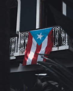 A Puerto Rican flag hangs from a wrought iron balcony, with intricate designs. The backdrop includes dark walls and hints of surrounding foliage creating a shadowy foreground.