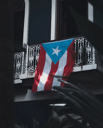 A Puerto Rican flag hangs from a wrought iron balcony, with intricate designs. The backdrop includes dark walls and hints of surrounding foliage creating a shadowy foreground.