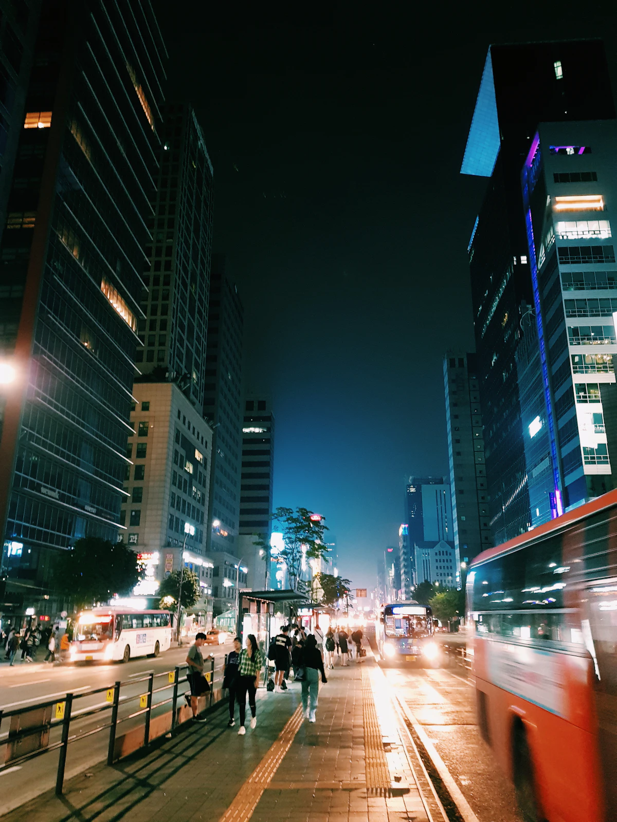 Wide sidewalk between glass office towers at night, pedestrians and a passing bus
