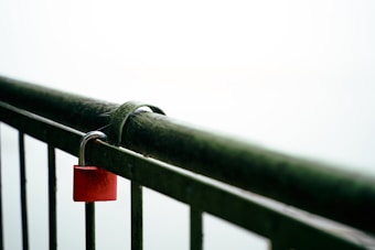 A red padlock is secured onto a metal railing, which appears to be slightly weathered and covered in moss. The background is a soft, light blur, suggesting a foggy or overcast day.