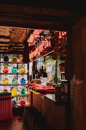 A dimly lit restaurant interior with a neon sign and multiple colorful glass bottles on a shelf. The countertop is lined with red plates and there are hanging pendant lights overhead. Natural light filters in through a window, and there's brickwork along the counter.