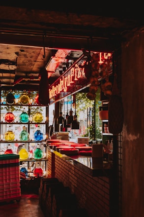 A dimly lit restaurant interior with a neon sign and multiple colorful glass bottles on a shelf. The countertop is lined with red plates and there are hanging pendant lights overhead. Natural light filters in through a window, and there's brickwork along the counter.