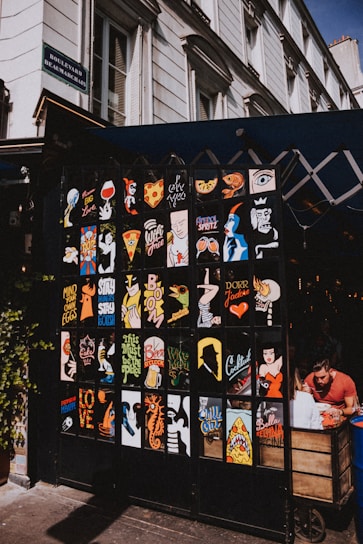 A vibrant display of pop art style posters with various colorful designs and phrases adorns the front of a small shop or café. The posters exhibit a mix of graphics including abstract faces, food items like pizza, and beverages. The setting is on a city street with the building marked 'Boulevard Beaumarchais'. To the right, a person is seated at a wooden cart, seemingly engrossed in their meal or activity.