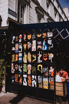 A vibrant display of pop art style posters with various colorful designs and phrases adorns the front of a small shop or café. The posters exhibit a mix of graphics including abstract faces, food items like pizza, and beverages. The setting is on a city street with the building marked 'Boulevard Beaumarchais'. To the right, a person is seated at a wooden cart, seemingly engrossed in their meal or activity.