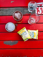 A top-down view of a wooden table painted in vibrant red, featuring two glasses of beer with frothy tops, an empty glass, and a collection of glasses with water. There are two packets of crisps named 'Scampi Fries' placed next to the drinks.