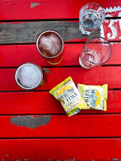 A top-down view of a wooden table painted in vibrant red, featuring two glasses of beer with frothy tops, an empty glass, and a collection of glasses with water. There are two packets of crisps named 'Scampi Fries' placed next to the drinks.