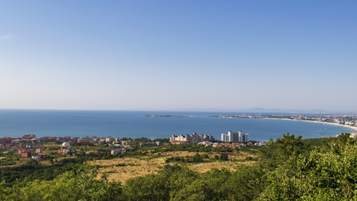 A coastal landscape features a vast expanse of blue ocean meeting the horizon under a clear sky. In the foreground, there's a mix of green vegetation, open fields, and a cluster of buildings, including some taller structures near the shore. The coastline curves gently, with visible distant hills and a clear beach.