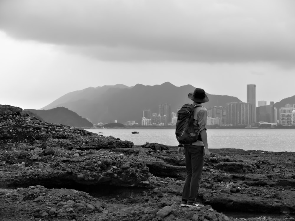 grayscale photography of man standing near sea viewing city with high-rise buildings