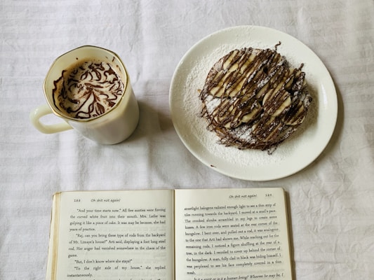 A cozy setup featuring a white mug filled with intricately designed coffee foam on the left. To the right is a plate with a dessert topped with sliced bananas, drizzled with chocolate syrup, and dusted with powdered sugar. An open book lies at the bottom of the image on a soft, white fabric background.