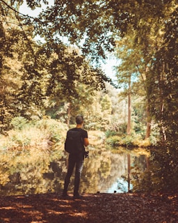 A person wearing a backpack stands near the edge of a calm body of water, surrounded by lush trees and foliage. The sunlight filters through the leaves, casting dappled shadows on the ground and the water's surface. The tranquil setting evokes a sense of peaceful solitude in nature.