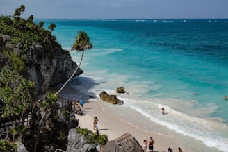 A scenic beach with turquoise waters meets white sand, bordered by rocky cliffs with green vegetation. Several people enjoy the water and walk along the beach, while others stand on a wooden walkway surrounded by tropical plants.