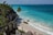 A scenic beach with turquoise waters meets white sand, bordered by rocky cliffs with green vegetation. Several people enjoy the water and walk along the beach, while others stand on a wooden walkway surrounded by tropical plants.