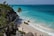 A scenic beach with turquoise waters meets white sand, bordered by rocky cliffs with green vegetation. Several people enjoy the water and walk along the beach, while others stand on a wooden walkway surrounded by tropical plants.