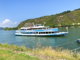 A two-level passenger boat traveling on a river. The boat is white with blue accents, featuring a logo and some text. Passengers are visible on the upper deck, enjoying the sunny weather. In the background, there are green hills with sparse trees, and part of the shoreline is visible in the foreground.