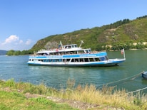 A two-level passenger boat traveling on a river. The boat is white with blue accents, featuring a logo and some text. Passengers are visible on the upper deck, enjoying the sunny weather. In the background, there are green hills with sparse trees, and part of the shoreline is visible in the foreground.