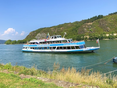 A two-level passenger boat traveling on a river. The boat is white with blue accents, featuring a logo and some text. Passengers are visible on the upper deck, enjoying the sunny weather. In the background, there are green hills with sparse trees, and part of the shoreline is visible in the foreground.