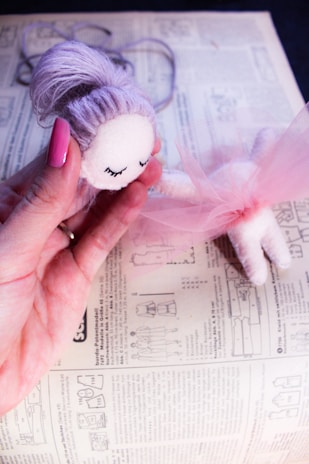 A hand holding a handmade doll with a soft, white face and closed eyes, paired with lavender yarn hair styled in a bun. The doll is wearing a pink tulle skirt and is positioned above a sewing pattern laid out on an instructional book.