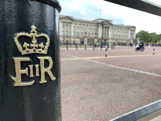 A close-up of a black metal post features the monogram 'E II R' with a crown emblem above. In the background, a grand building is visible, suggesting an important or historical site. The scene includes a road and a motorcyclist nearby.