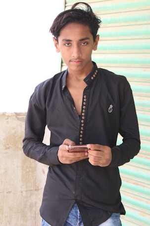 A person with short hair is wearing a black button-up shirt with a decorative emblem and holding a smartphone. The person is standing in front of a textured concrete wall and a closed, green metal roller shutter.