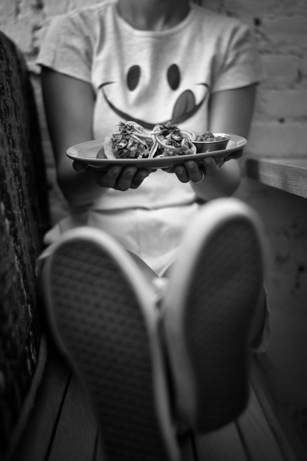 grayscale photography of woman holding plate with food while sitting and leaning near wall