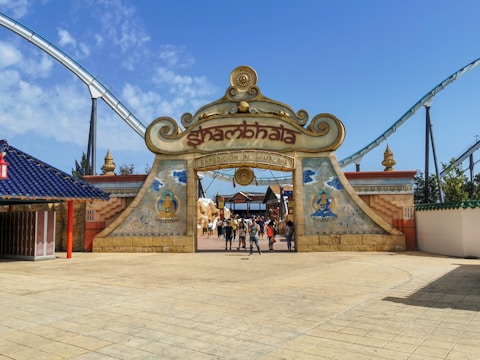 An entrance gate with an ornate design featuring the word 'Shambhala' and decorative elements including Buddhist symbols and art. People are walking through the gate, and a rollercoaster track is visible in the background.
