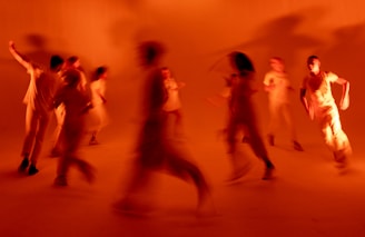 Blurred figures appear to be moving dynamically in a warm, orange-lit space. The image conveys motion and energy with the figures' movements being captured in a state of motion blur.
