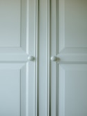 A close-up of a light-colored cabinet door featuring raised panels and two round white knobs. The cabinet's surface has a smooth finish with slight shadows highlighting the contours of the panels.