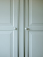 A close-up of a light-colored cabinet door featuring raised panels and two round white knobs. The cabinet's surface has a smooth finish with slight shadows highlighting the contours of the panels.