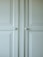 A close-up of a light-colored cabinet door featuring raised panels and two round white knobs. The cabinet's surface has a smooth finish with slight shadows highlighting the contours of the panels.