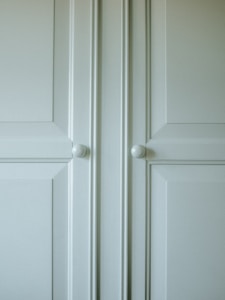 A close-up of a light-colored cabinet door featuring raised panels and two round white knobs. The cabinet's surface has a smooth finish with slight shadows highlighting the contours of the panels.